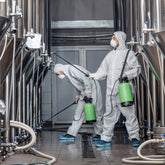 <Two men dressed in protective white overalls, wearing safety boots, face masks and gloves, cleaning the outside of manufacturing mixing tanks with spray hoses from a green chemical container. Credit> AdobeStock_376451842