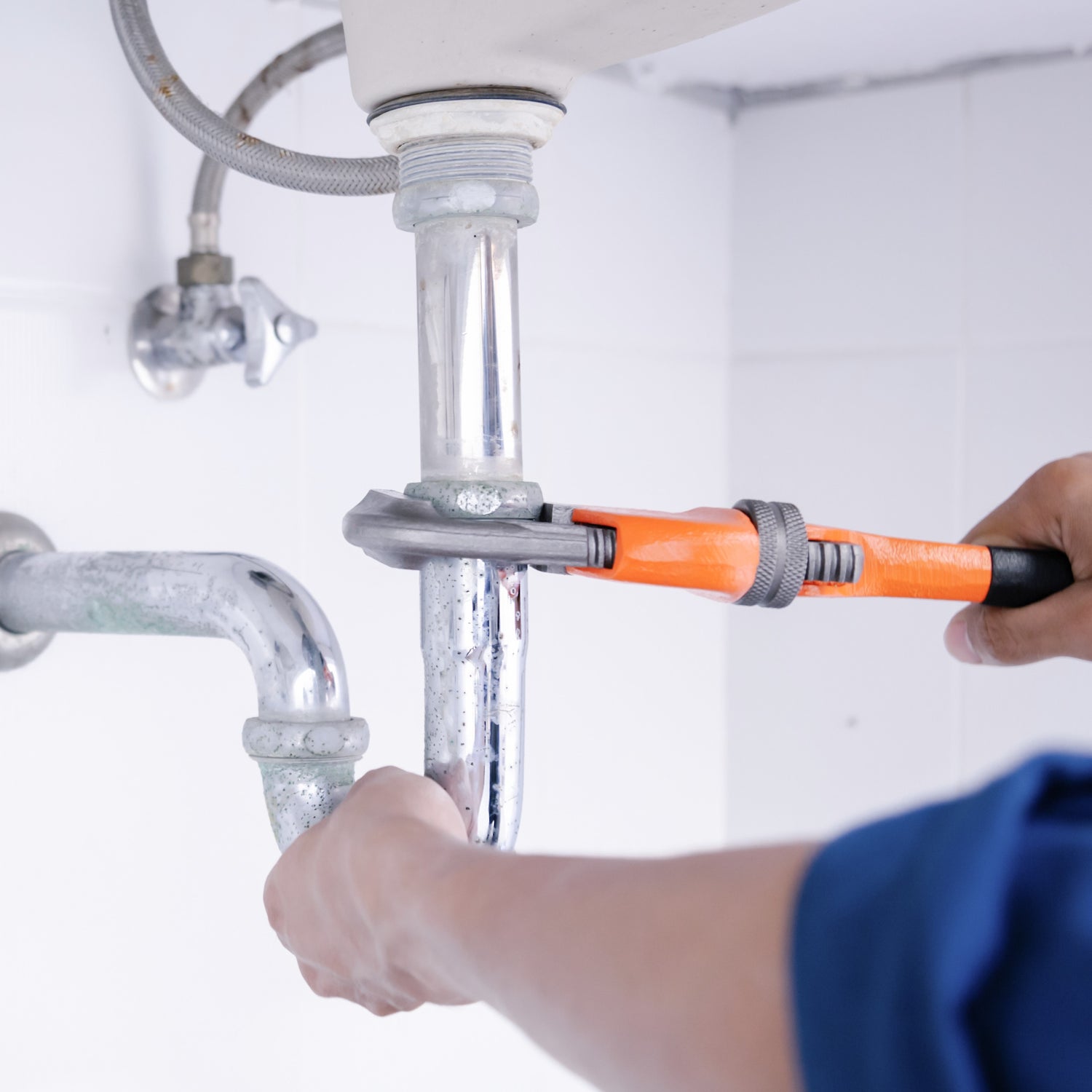 &lt;Engineer in blue overall fixing the leaking chrome waste plumbing with an orange adjustable wrench underneath an employee white handwash sink. Credit&gt; AdobeStock_402968661