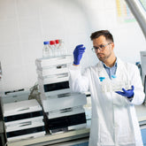 <Male QC Analyst in front of an HPLC looking at a sample in a flask performing the lab tests to determine identity, purity, strength, and composition of the dietary supplement. Credit> AdobeStock_403421068