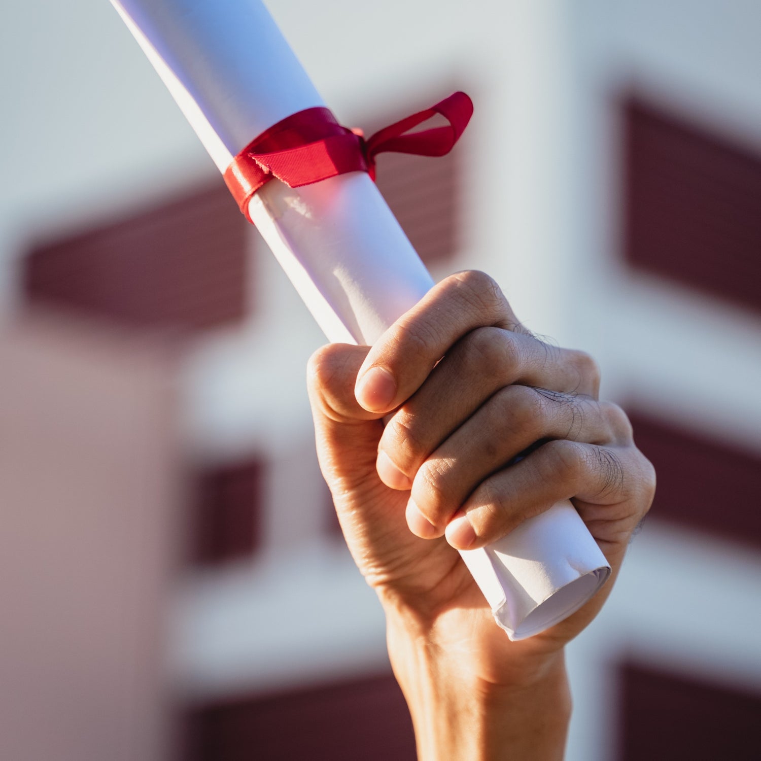 &lt;Hand holding up a rolled up diploma with a red ribbon in celebration of qualifying, with a purple and white blurred college building behind. Credit&gt; AdobeStock_406407277