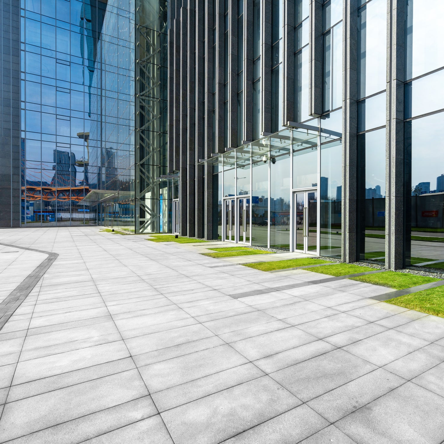 &lt;Stone paved area with symmetric patterns in front of a modern glass L shaped office building with small green areas. Credit&gt; AdobeStock_414022077