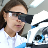 <Female laboratory analyst wearing white coat, blue gloves, and safety specs looking through the binocular eyepiece of a white microscope. Credit> AdobeStock_427358393