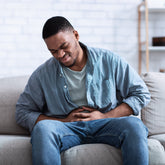 <Black male employee, wearing light blue denim jeans and shirt, sitting on grey sofa at home in pain with a stomach upset. White brick wall and storage shelves behind him. Credit> AdobeStock_429222244