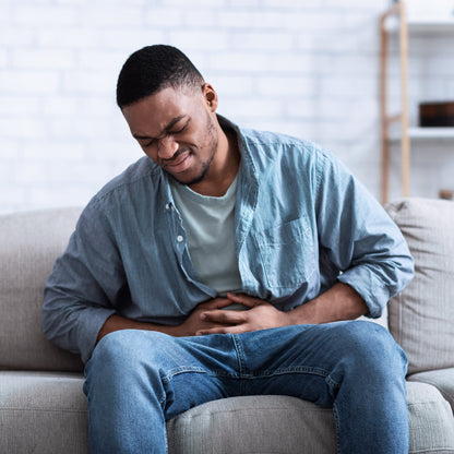 &lt;Black male employee, wearing light blue denim jeans and shirt, sitting on grey sofa at home in pain with a stomach upset. White brick wall and storage shelves behind him. Credit&gt; AdobeStock_429222244