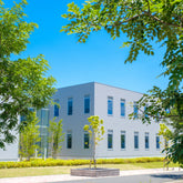 <Outside of a grey metal and glass drug facility on a bright sunny day with well kept green spaces, pathways and trees. Credit> AdobeStock_437323910