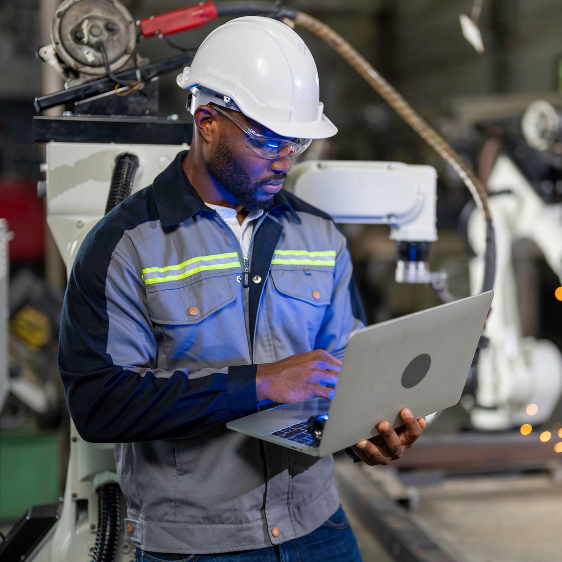 &lt;Maintenance Enginneer wearing blue overall, white hardhat and safety glasses analyzing a white robotic arm packing unit with a laptop computer. Credit&gt; AdobeStock_438287786