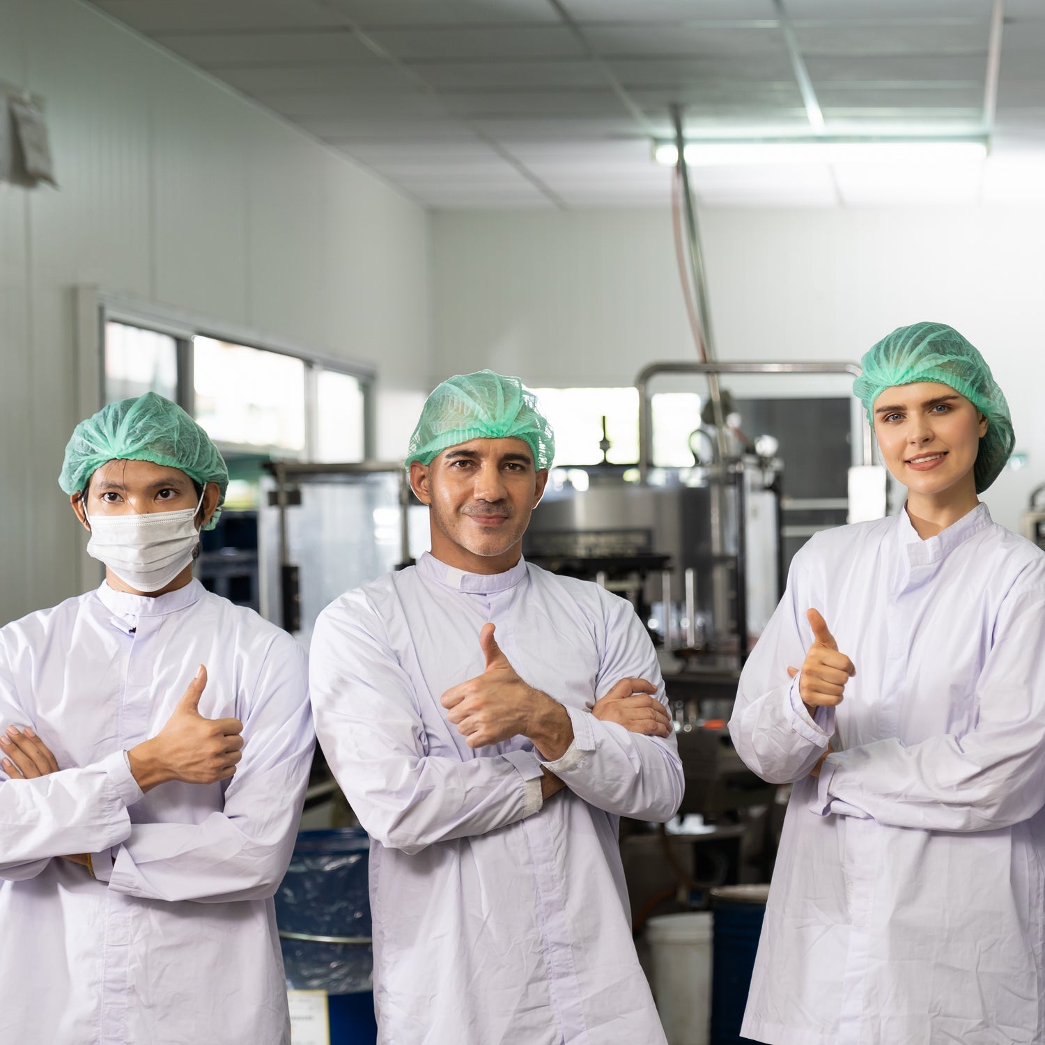 &lt;Three smiling employees near a filling line, two male and one female, giving a thumbs up signal for wearing the correct white coat and blue hairnet. Credit&gt; AdobeStock_454159606