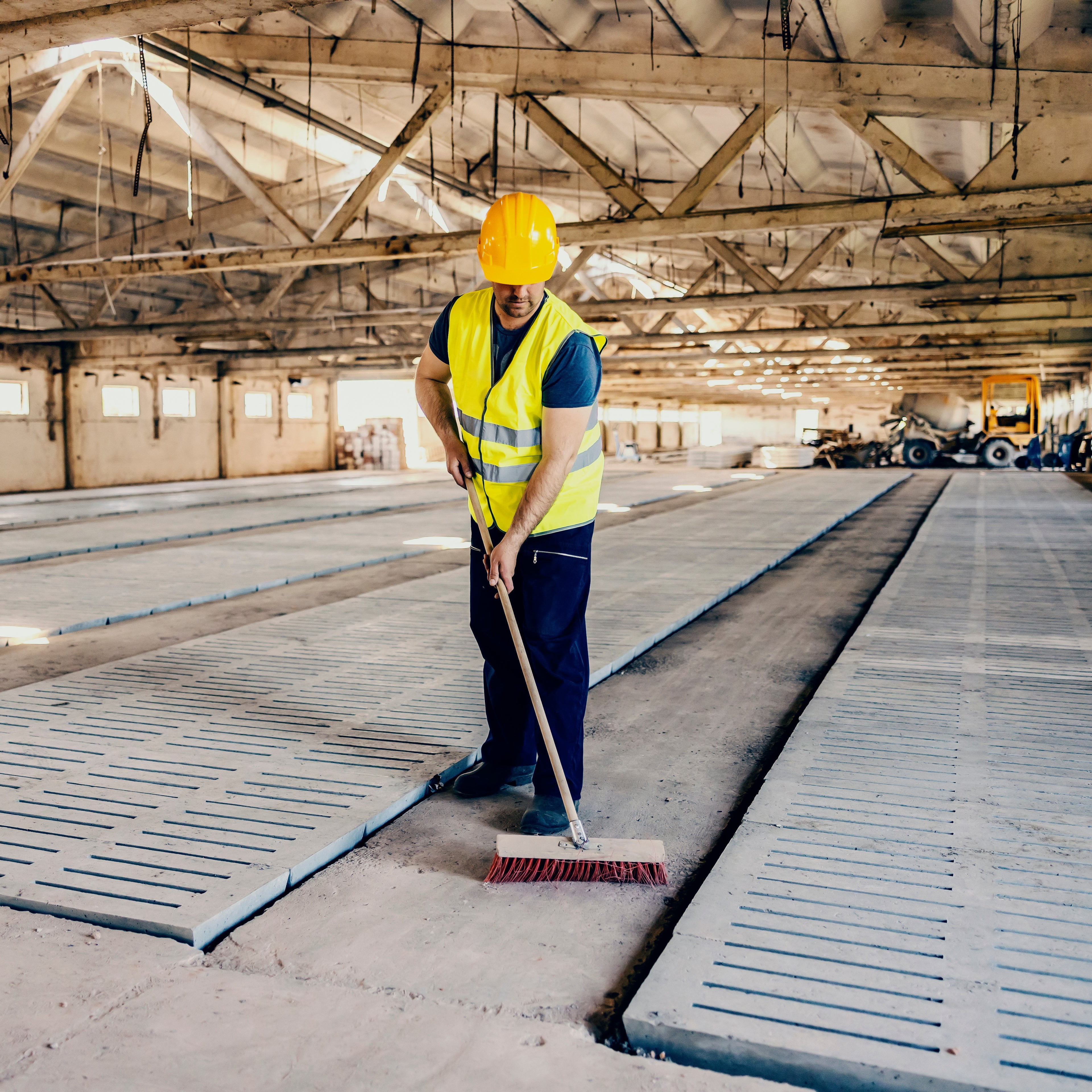 &lt;Man wearing dark blue trousers, safety boots and a yellow fluorescent jacket and hardhat, sweeping the concrete floor of a large warehouse with a brush to remove any dust. Credit&gt; AdobeStock_485391655