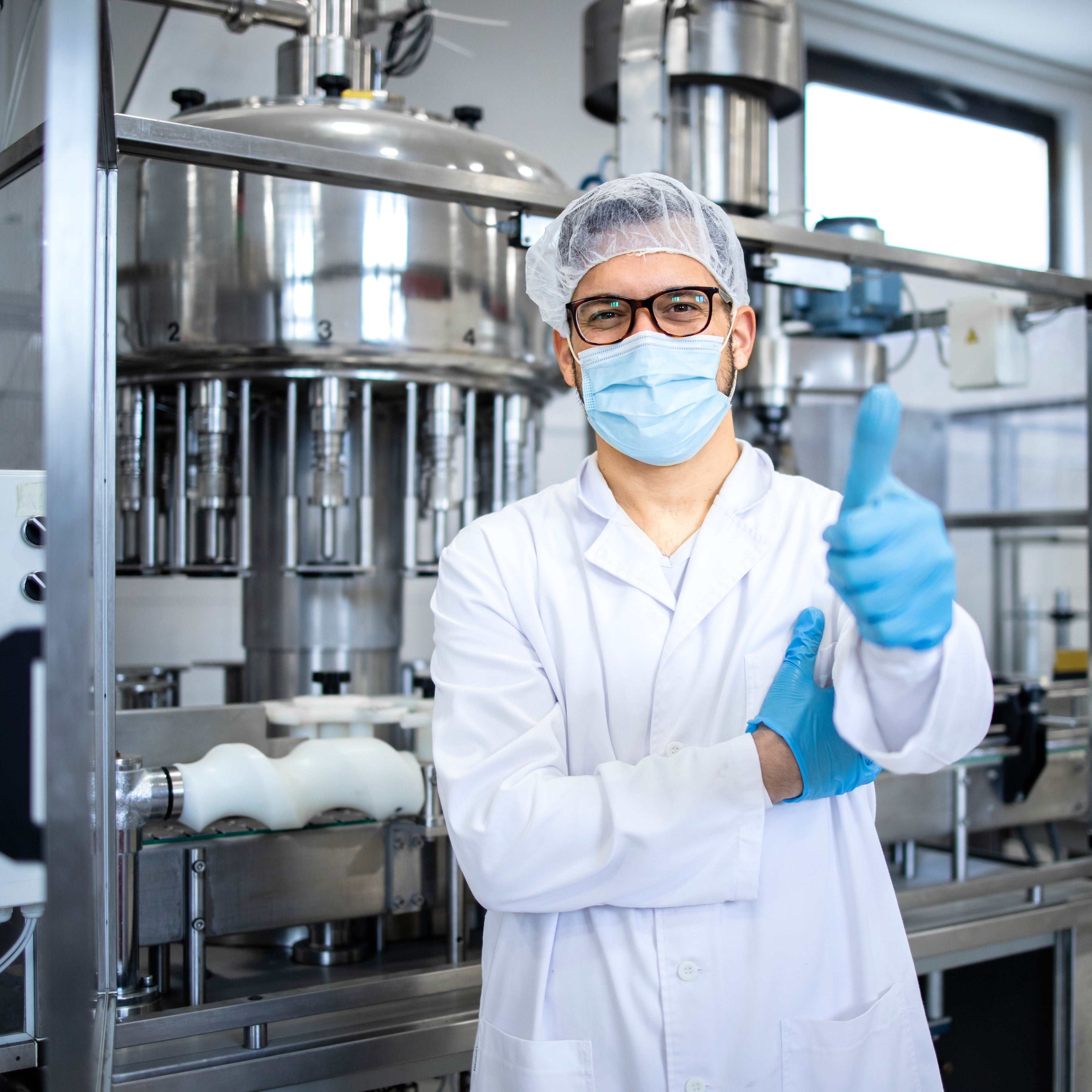 &lt;White, smiling male production operator with glasses wearing a white coat, hairnet and face mask with blue gloves giving the thumbs up stood in front of a tablet filling machine. Credit&gt; AdobeStock_487453879