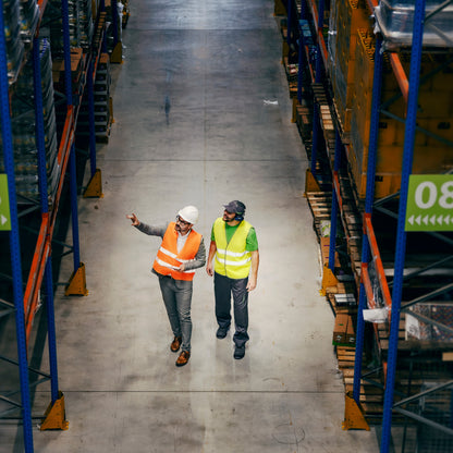 &lt;Looking down at two male employees walking down an aisle between racking in a warehouse, one wearing orange and white hard hat pointing up, one wearing yellow and blue hard hard looking. Credit&gt; AdobeStock_489525726