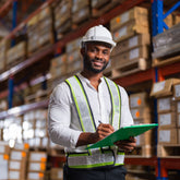 <Male, smiling warehouse operator, wearing fluorescent vest and white hard hat, walking down an aisle in a warehouse writing on a clipboard. Credit> AdobeStock_505465613