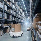 <Automated robotic pallet truck on concrete floor, delivering a large box to the racking of a large warehouse in sections five pallet spaces high and over twenty pallet spaces wide. Credit> AdobeStock_507641871