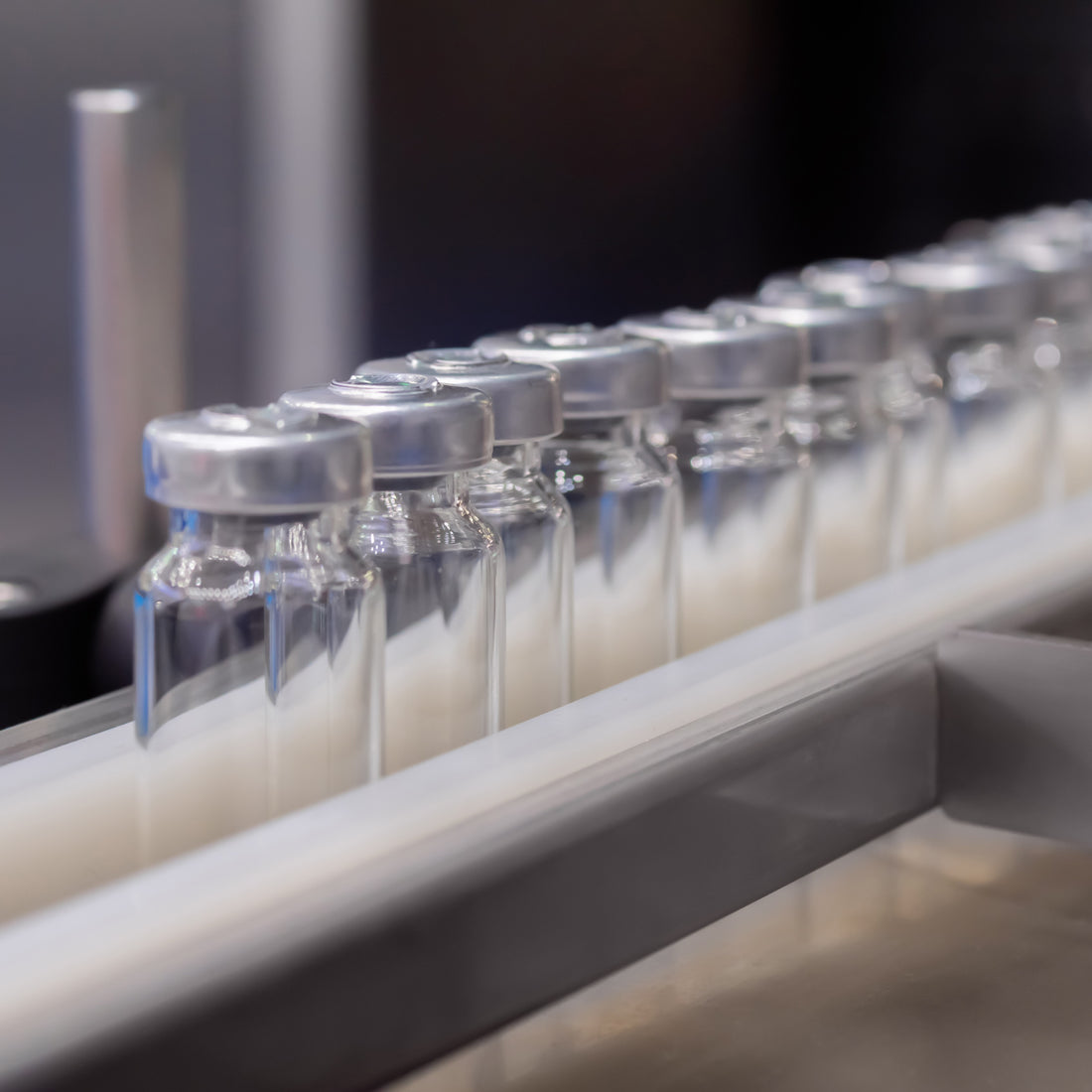 &lt;Row of glass vaccine vials with metal caps passing down a conveyor belt with guardrails keeping them in place to be filled. Credit&gt; AdobeStock_513373066