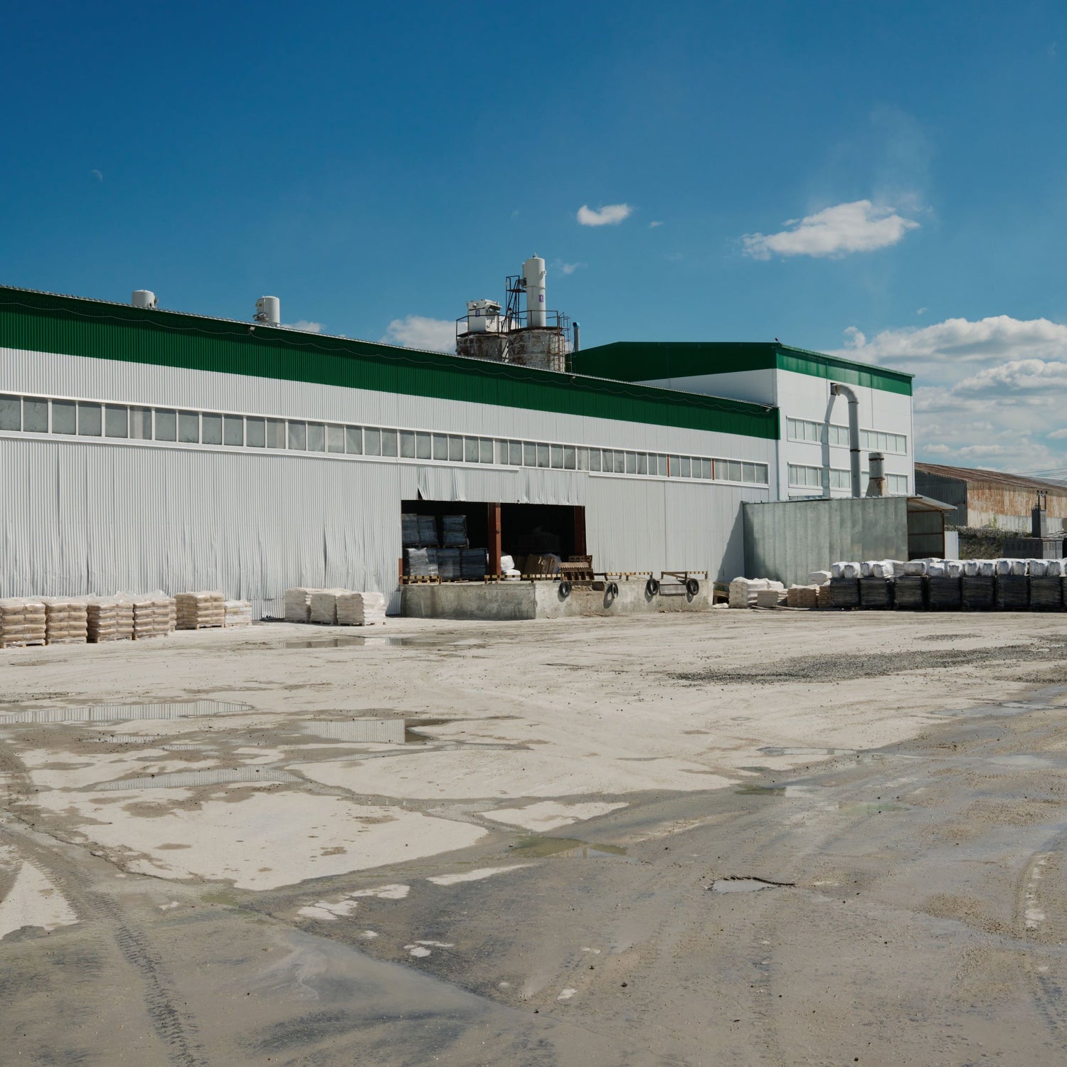 &lt;Concrete yard, in sunshine after rain, of a grey and green striped corrugated metal dietary supplement production  building taking in pallet deliveries of materials through a shutter door. Credit&gt; AdobeStock_525171795