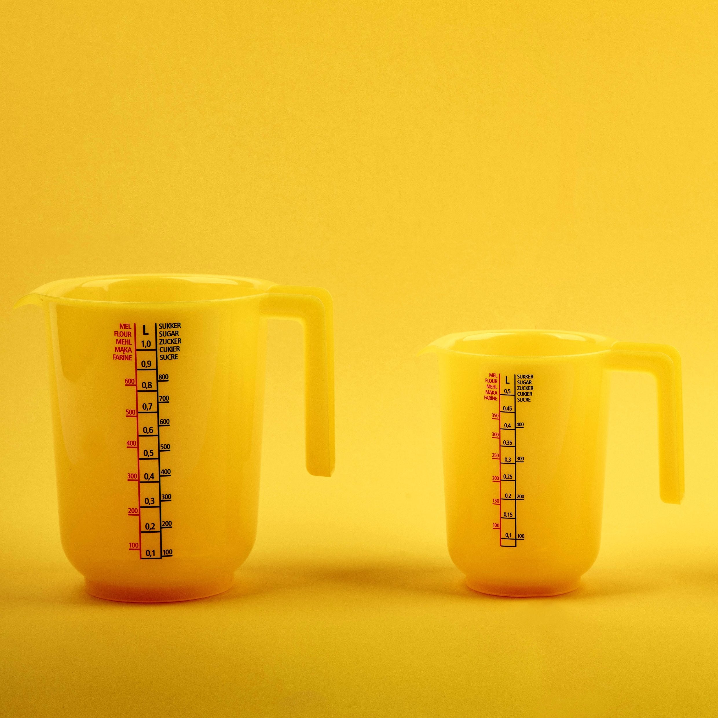 &lt;One large and one medium plastic jug, used for measuring the volume of liquid supplement ingredients, sitting on a smooth surface with a bright yellow lit background. Credit&gt; AdobeStock_544731939