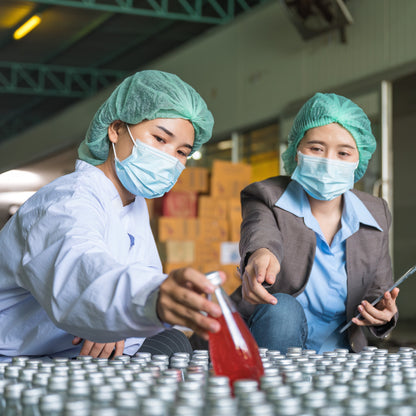 &lt;Two female Quality Control officers,  wearing blue hairnets and face masks, one with a clipboard pointing at and the other inspecting 1 from over 100 bottles of red, liquid dietary supplement with a  silver metal cap in a production area. Credit&gt; AdobeStock_551742643