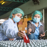 <Female, senior quality manager in production pointing to a bottle of red liquid with silver cap that she would like to inspect from hundreds, with a female production operator selecting it for her. Credit> AdobeStock_551742643