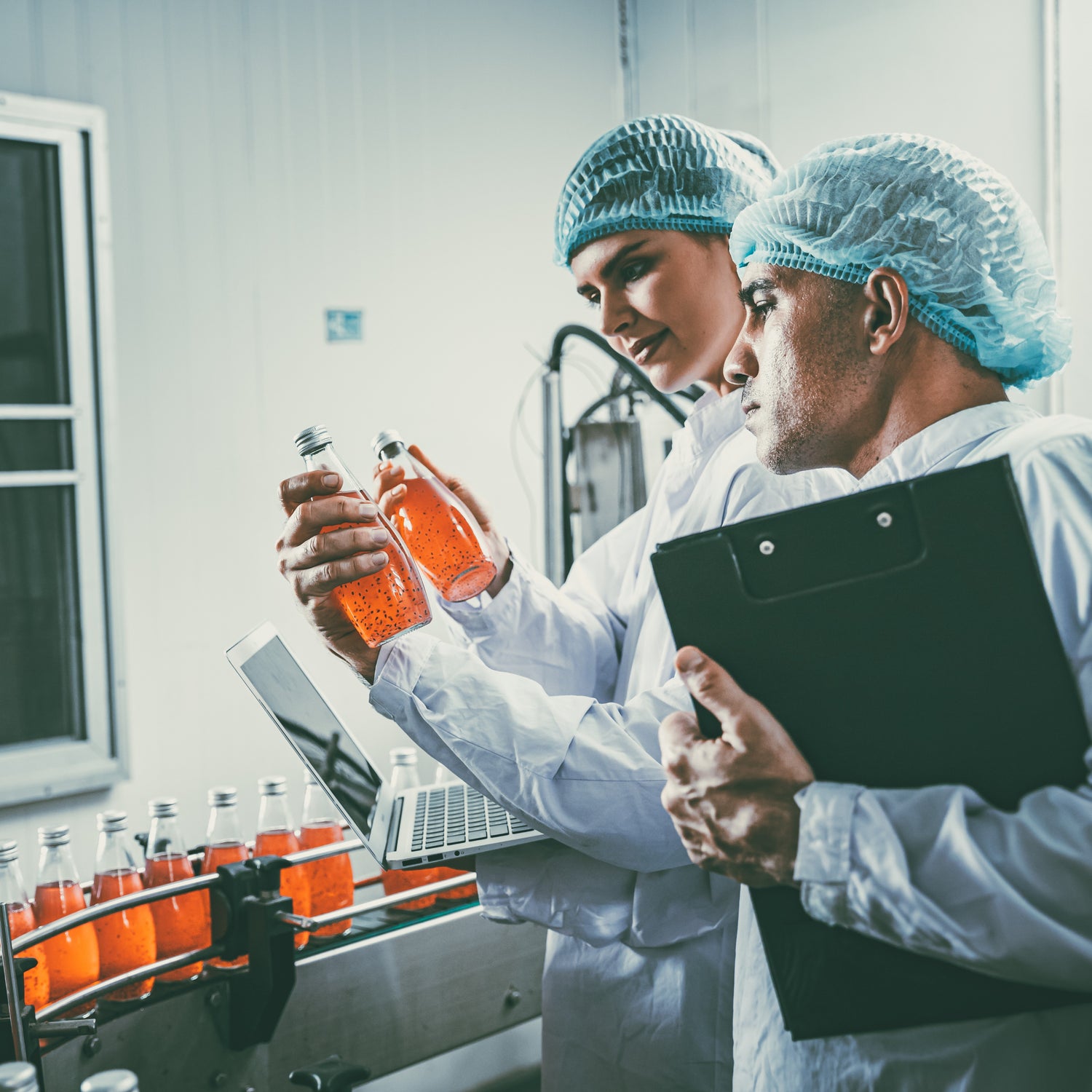 &lt;Two Quality Officers, one male with a clipboard, the other female with a laptop, both wearing white coats and blue hairnets, inspecting bottles of orange liquid running down a production line conveyor belt,. Credit&gt; AdobeStock_552543492