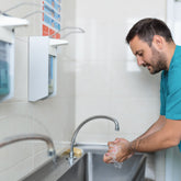 <Bearded man wearing green t-shirt washing his hands using one of the liquid soap wall dispensers hung on a white, smooth tiled wall above a long stainless steel sink. Credit> AdobeStock_562119066