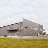 <Looking across a well kept grass area and tarmac yard to a grey metal drug facility with a row of first floor windows and a spiral fire escape from the roof. Credit> AdobeStock_564629963