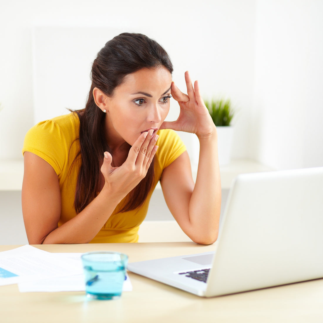 &lt;Female customer wearing a yellow short sleeve top at home in a minimalistic white living room, sat at a pale wooden table, blue glass of water, visibly shocked hand to mouth reading the response by e-mail to a complaint she made on a white laptop. Credit&gt; AdobeStock_78493419 