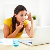 <Female customer wearing a yellow short sleeve top at home in a minimalistic white living room, sat at a pale wooden table, blue glass of water, visibly shocked hand to mouth reading the response by e-mail to a complaint she made on a white laptop. Credit> AdobeStock_78493419 