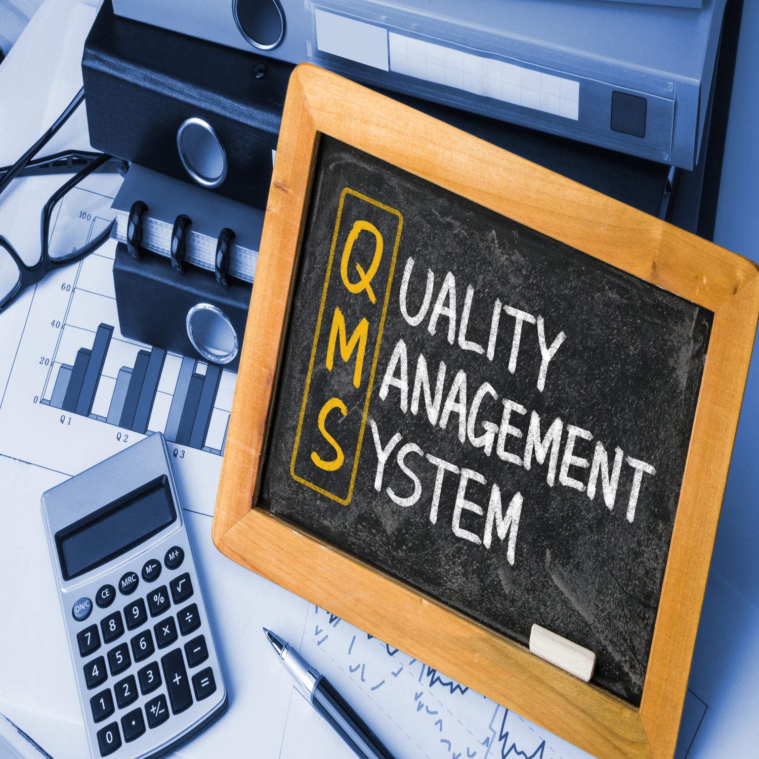 &lt;Small, wooden framed blackboard with Quality Management System written in white chalk, except for the letters QMS written and circled in yellow, sitting on a desk against some box files next to a calculator, pen and glasses. Credit&gt; AdobeStock_84985243