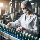 <Female production operator wearing white coat, hairnet and gloves inspecting green plastic bottles of a liquid dietary supplement with a blue label and silver metal cap on a production line conveyor belt. Credit> Freepik