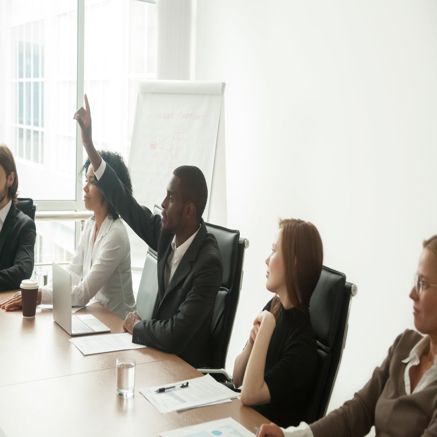 &lt;Five employees, three female, two male, in a training session in a white meeting room with wooden desk and flipchart, one of the males holding his hand up to ask a question. Credit&gt; 116558699 © Fizkes | Dreamstime.com