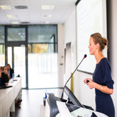 <Female representative from a supplier giving a presentation from a podium with microphone and pointer on a large screen to an audience in a white conference room. Credit> 38448112 © Lightpoet | Dreamstime.com